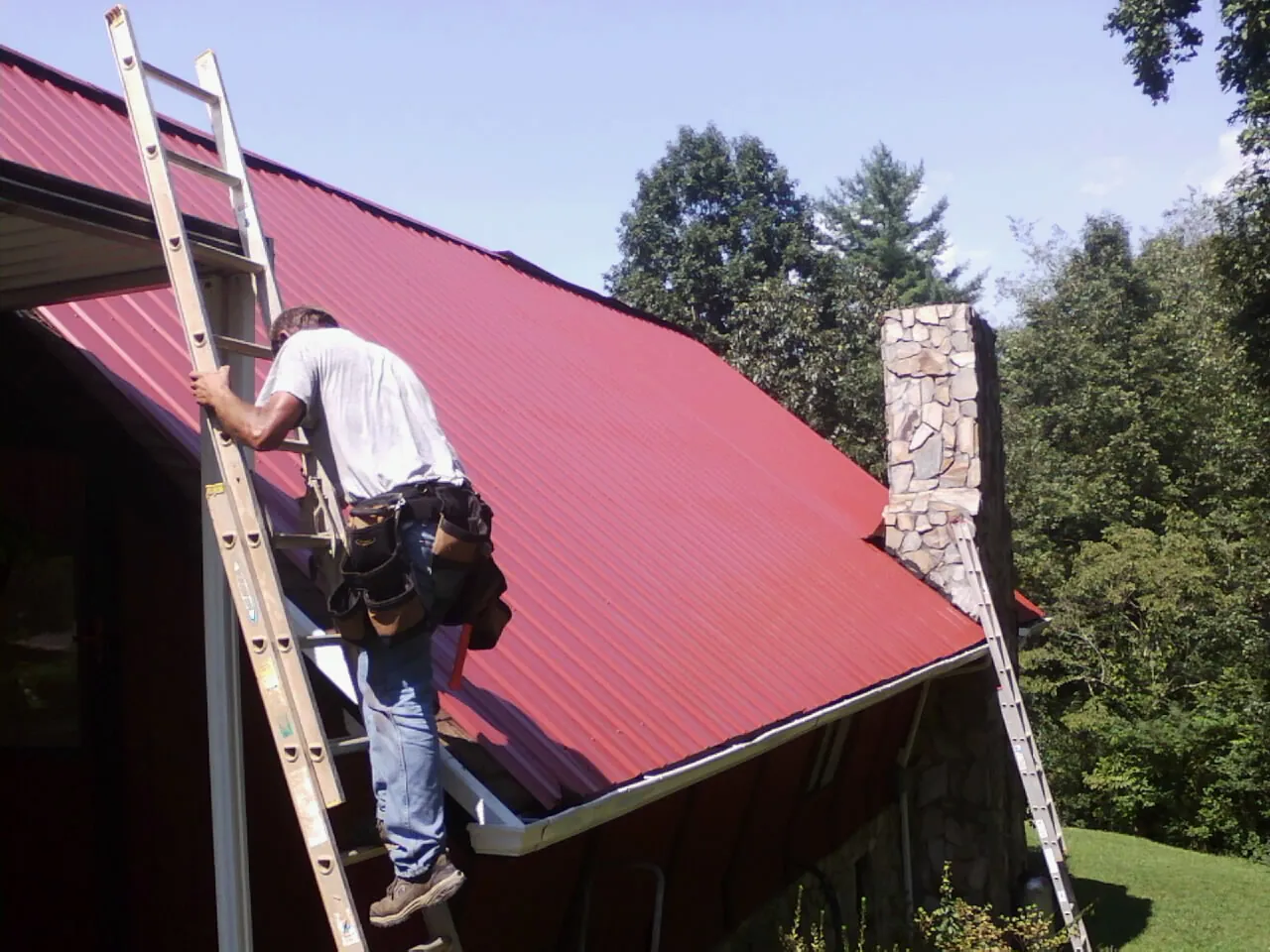 Roofer on a ladder beside a red metal roof and stone chimney — Soffit & Fascia Repair by Roofing Brothers in Mc Causland
